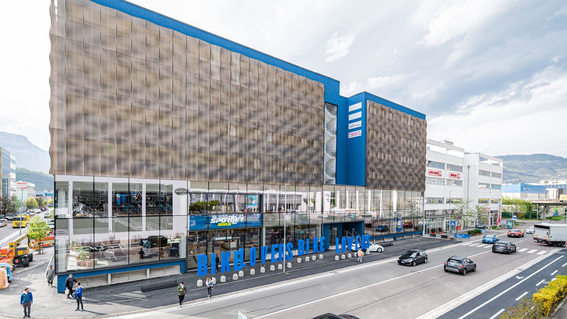 Modernes Firmengebäude von KRAISS mit Fahrrad- und Sportartikeln im Schaufenster, Blick auf die Straße mit Autos und Fußgängern, bei bewölktem Himmel.