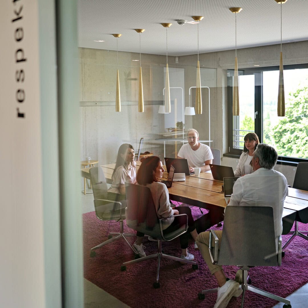 Konferenzraum mit fünf Personen, moderner Beleuchtung, große Fenster mit Blick auf Bäume, KRAISS Logo sichtbar an der Tür, stilvolle Einrichtung.