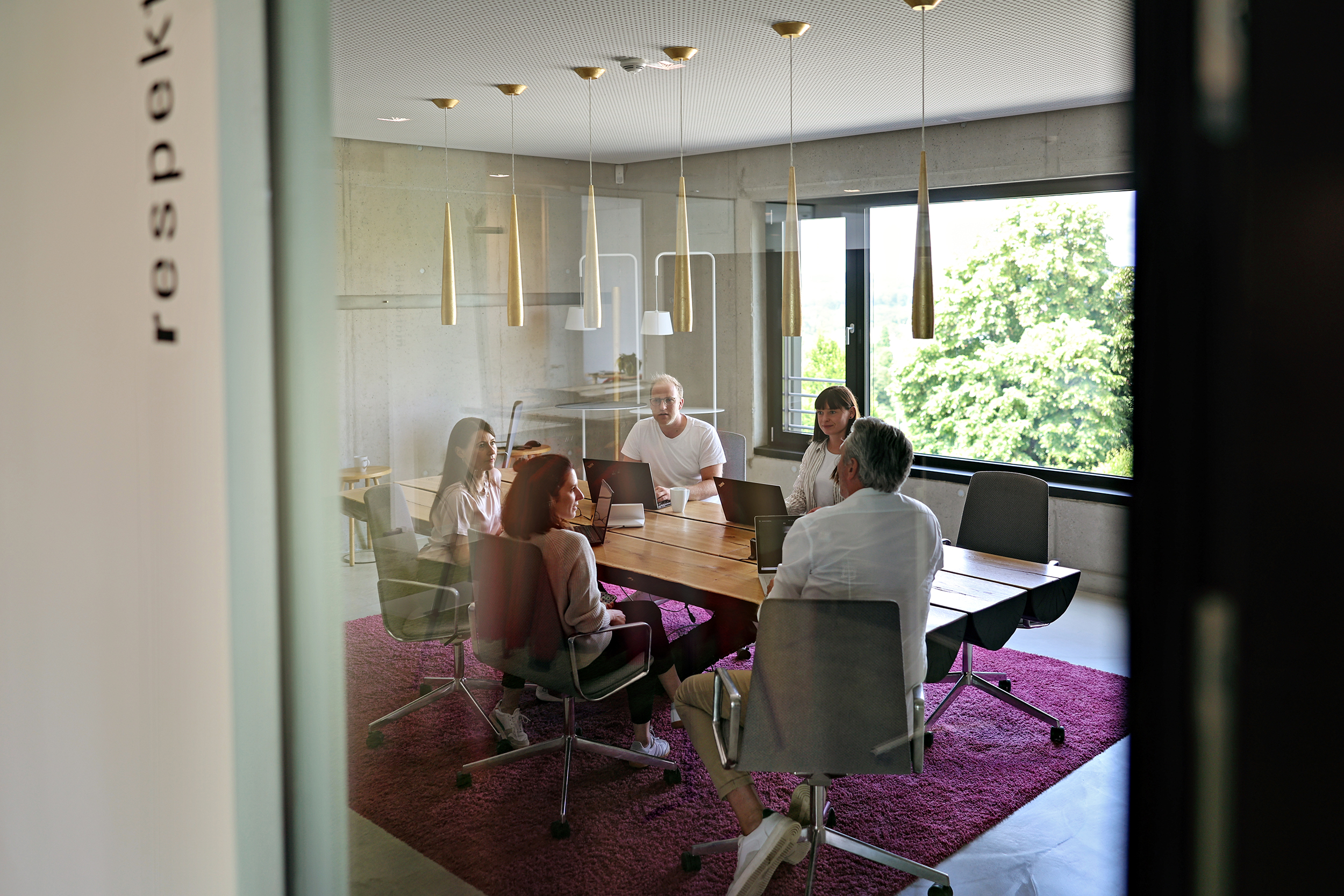Konferenzraum mit fünf Personen, moderner Beleuchtung, große Fenster mit Blick auf Bäume, KRAISS Logo sichtbar an der Tür, stilvolle Einrichtung.
