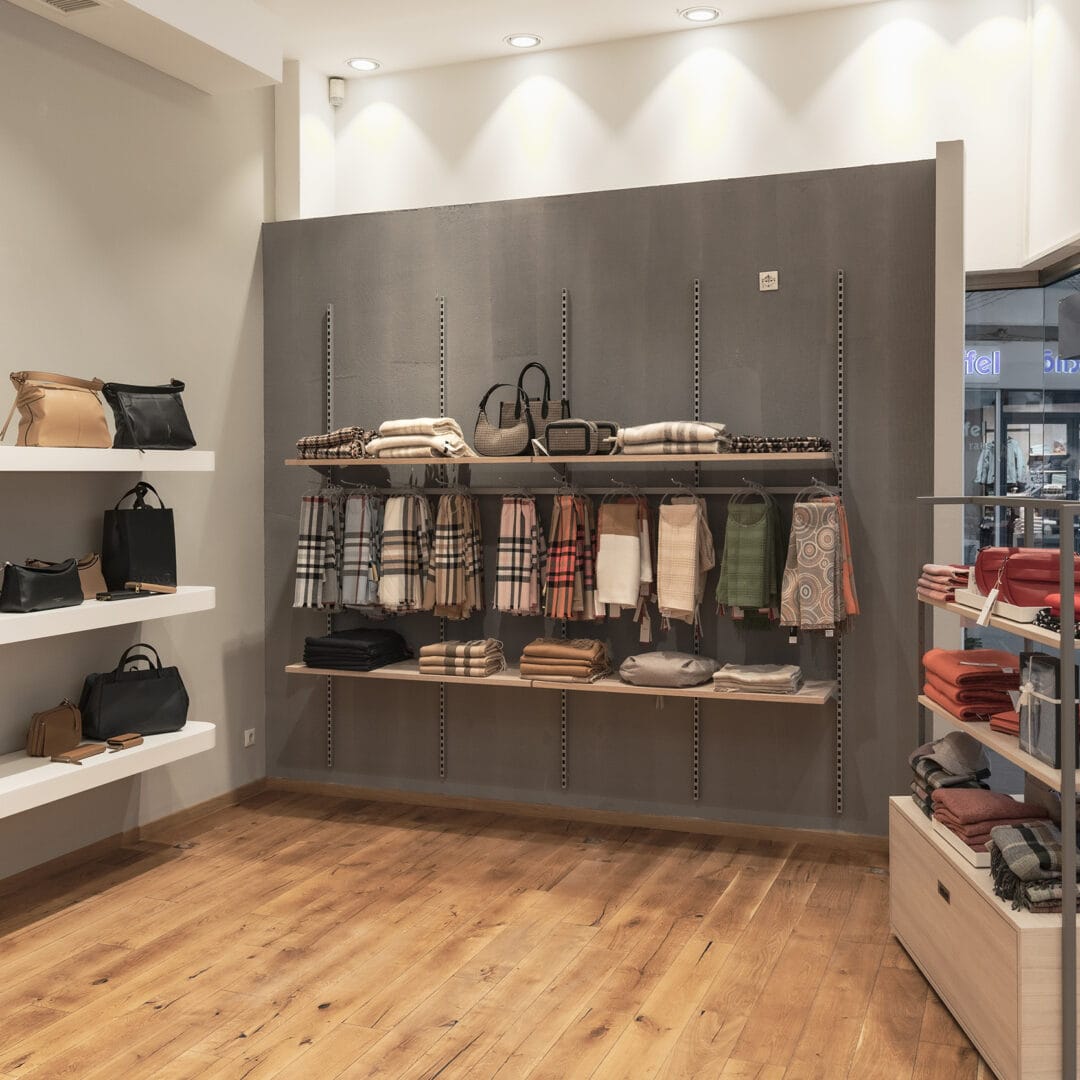 Innenansicht eines Ladenlokals mit Verkaufsregalen für Taschen, Schals und Accessoires, beleuchtet mit Deckenstrahlern.