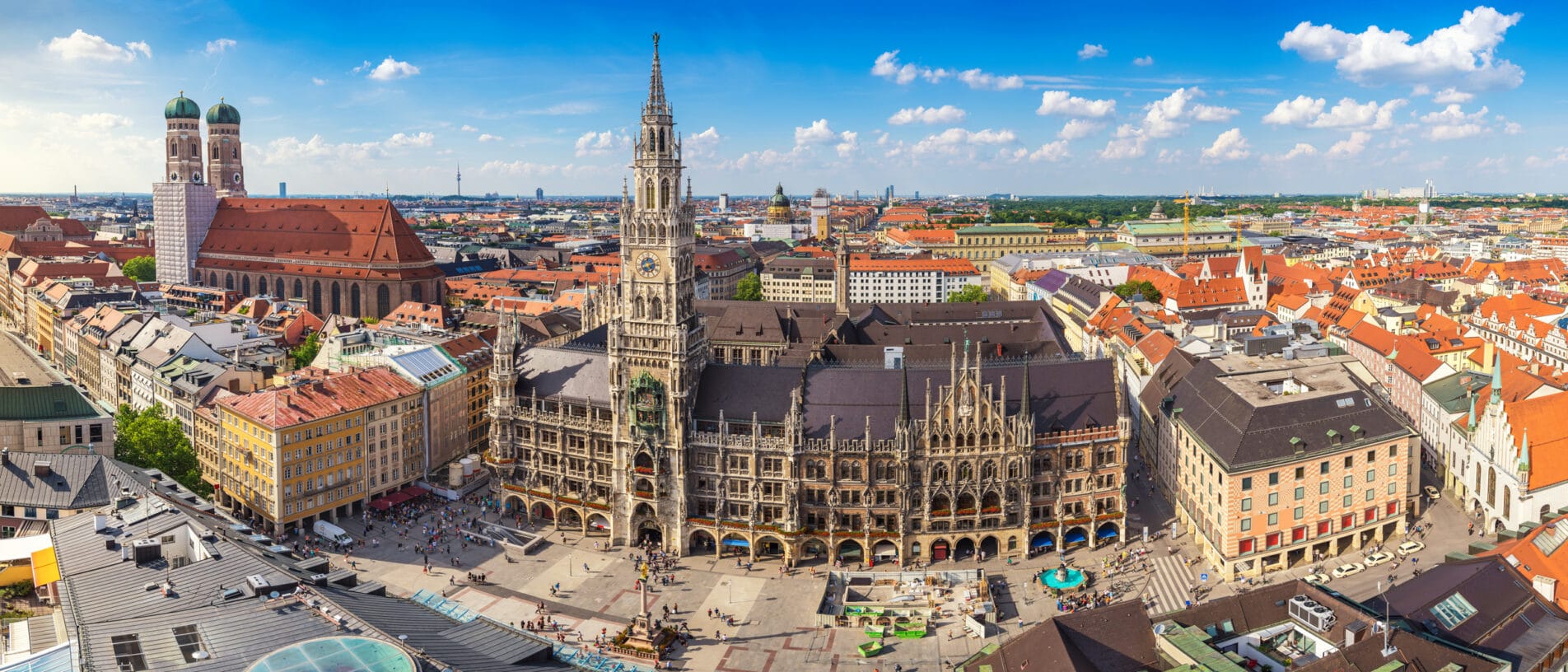 Panoramablick auf das historische Stadtbild Münchens mit dem Rathaus und der Frauenkirche bei sonnigem Himmel.