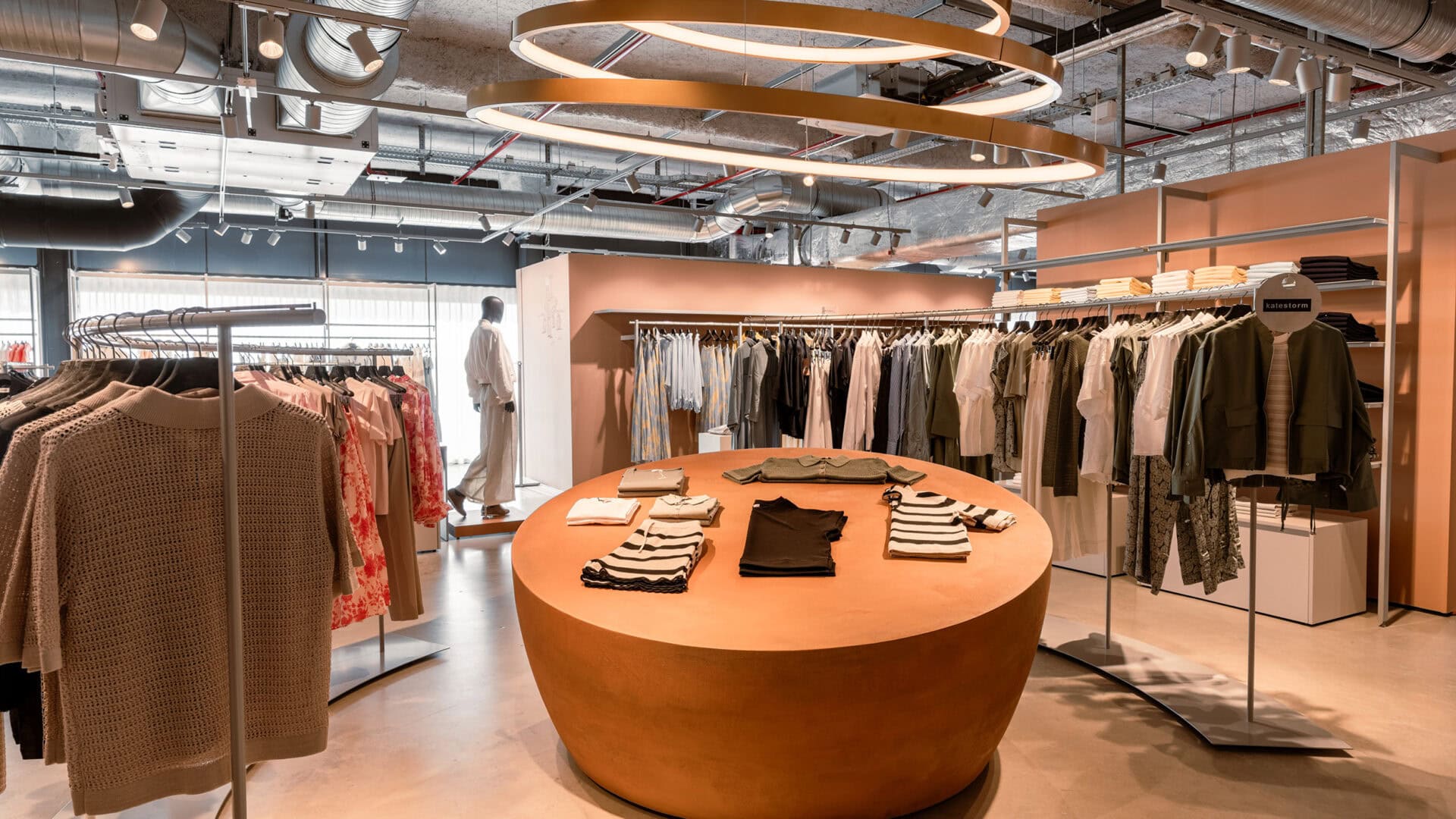 Moderne Modeboutique von KRAISS mit Kleidung auf Kleiderstangen und einem runden Holzdisplay in einem stilvollen, urbanen Ladenlokal.