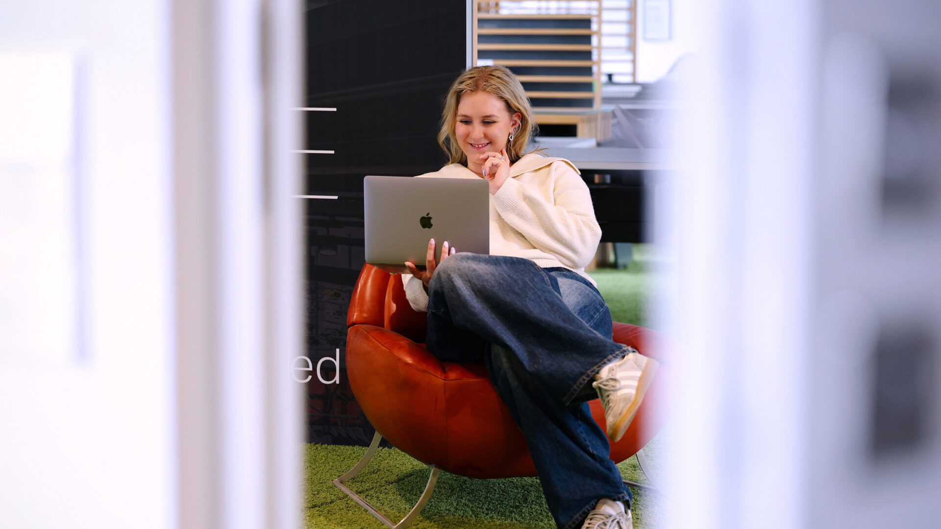 Junge Frau arbeitet entspannt mit Laptop in einem modernen Büro bei KRAISS, umgeben von innovativen Möbeln und Arbeitsbereichen.