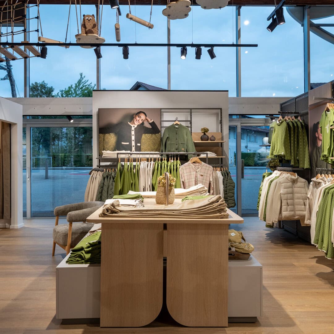 Innenansicht eines Modegeschäfts mit grüner Kleidung, Accessoires und großen Fenstern im Hintergrund.