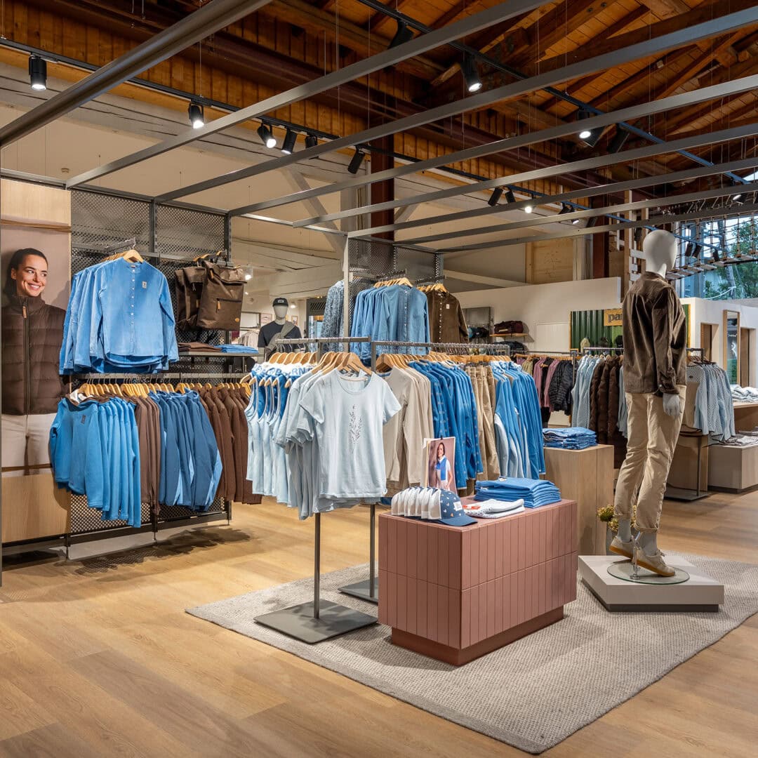Innenansicht eines Modegeschäfts mit Kleidung in Blau, auf Kleiderstangen und Regalen, mit Mannequins und Holzdecke.