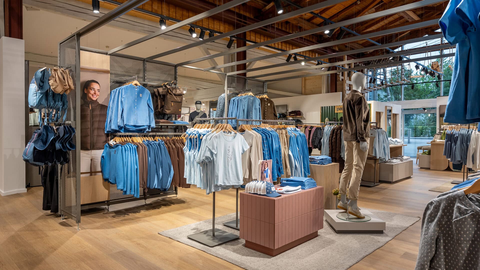 Innenansicht eines Modegeschäfts mit Kleidung in Blau, auf Kleiderstangen und Regalen, mit Mannequins und Holzdecke.