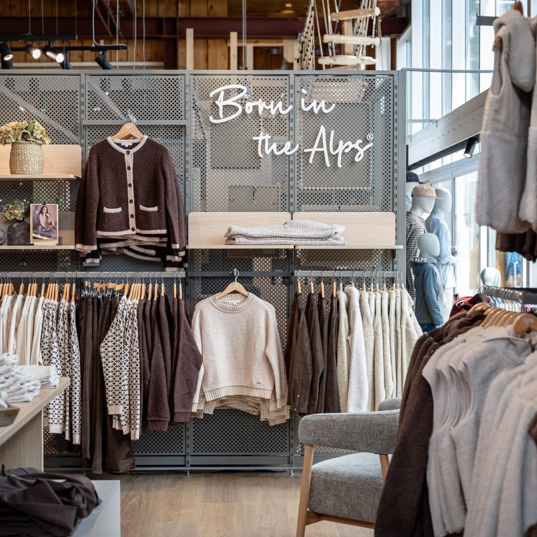Innenansicht eines Modegeschäfts mit Kleidung auf Hänge- und Regalböden, inklusive Jacken, Pullover und Accessoires.