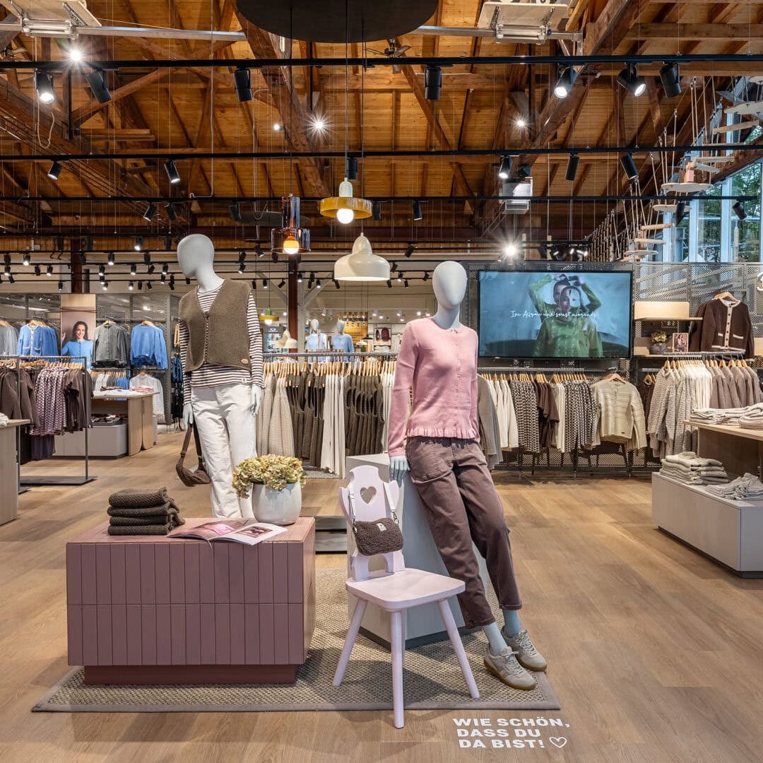 Innenansicht eines Modegeschäfts mit Mannequins, Kleidung und Dekorationen, Holzdecke und große Fenster.