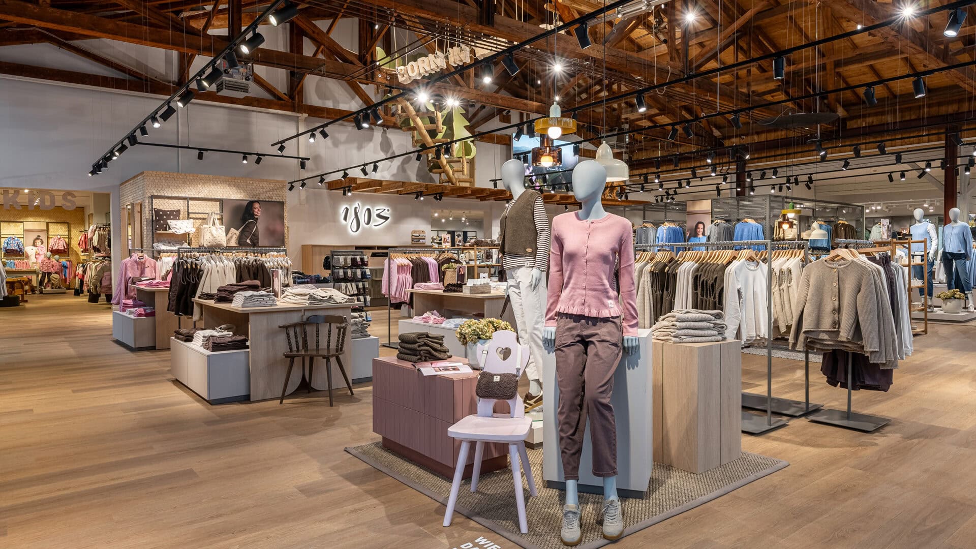 Innenansicht eines Modegeschäfts mit Mannequins, Kleidung und Dekorationen auf Holzfußboden und offenen Deckenstrukturen.