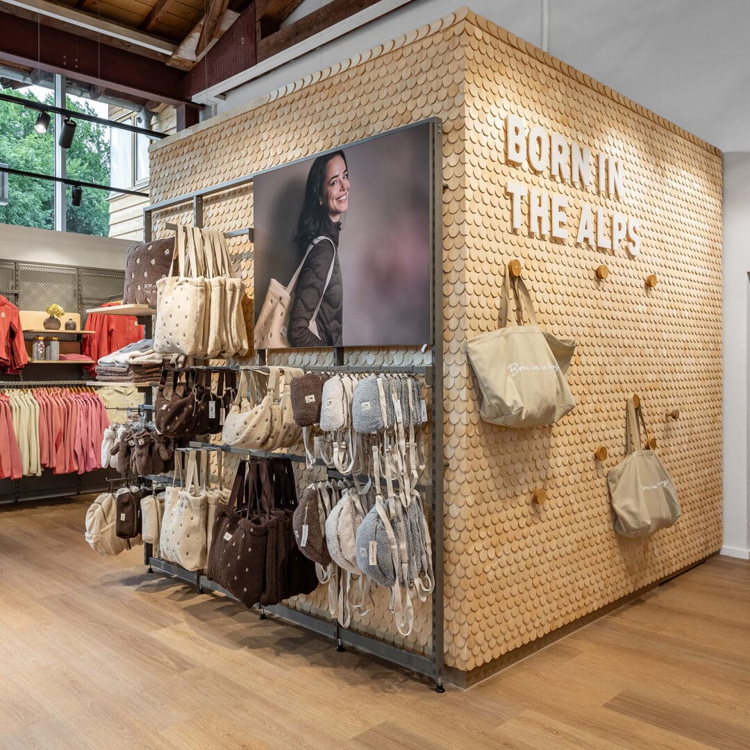 Verkaufstheke mit Taschen und Kleidung im 1803 Store Sonthofen, Holzfußboden, große Fenster, Markenlogo an Wand.
